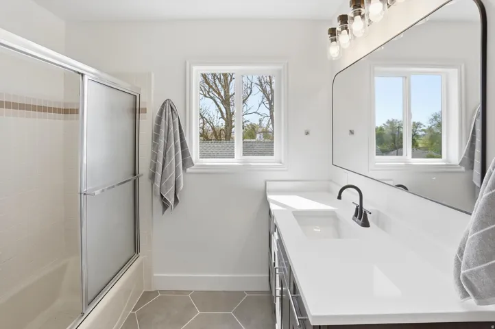 Bathroom featuring a shower-tub combination with sliding frosted glass doors and tile surround, a single vanity with a white countertop and dark cabinetry, a black framed arched mirror, and hexagonal floor tiling