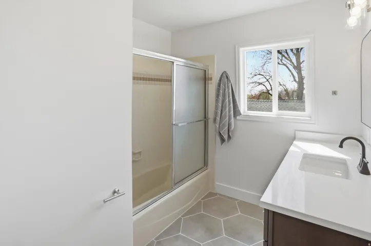 Bathroom featuring a tub/shower combination with sliding glass doors, a single vanity with a white countertop and undermount sink, hexagonal tile flooring, and a window providing natural light