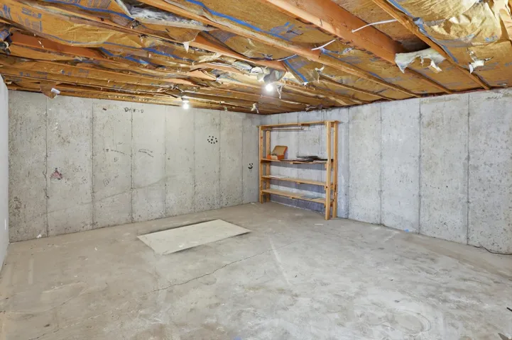 Unfinished basement space featuring concrete walls, concrete flooring, exposed ceiling joists, and a built-in wooden shelving unit