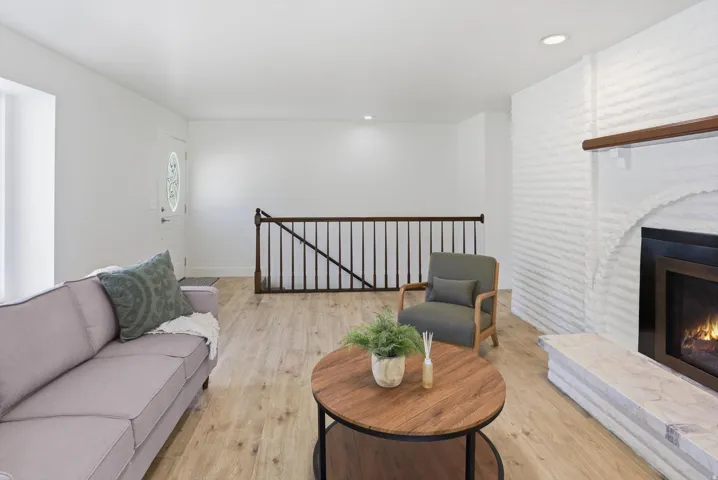 Living area featuring light wood-finish flooring, a white painted brick fireplace with a wood mantle and stone hearth, and a staircase with dark wood railing