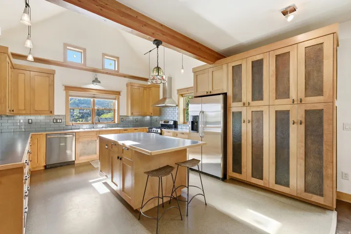 Kitchen featuring a vaulted ceiling with exposed wood beams, a central island with countertop, stainless steel appliances, and extensive cabinetry with textured panel inserts