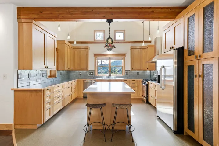 Spacious kitchen featuring light wood cabinetry, stainless steel appliances, a central island with countertop, subway tile backsplash, and exposed wooden beams