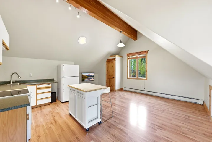 Vaulted ceiling with exposed wood beams, wood-finish flooring, recessed lighting, double-pane window with wood trim, and built-in kitchen cabinetry with a double basin sink