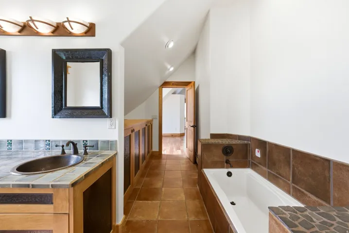 Bathroom featuring a built-in soaking tub with stone tile surround, a vanity with a copper sink and tile countertop, and wood-finish flooring