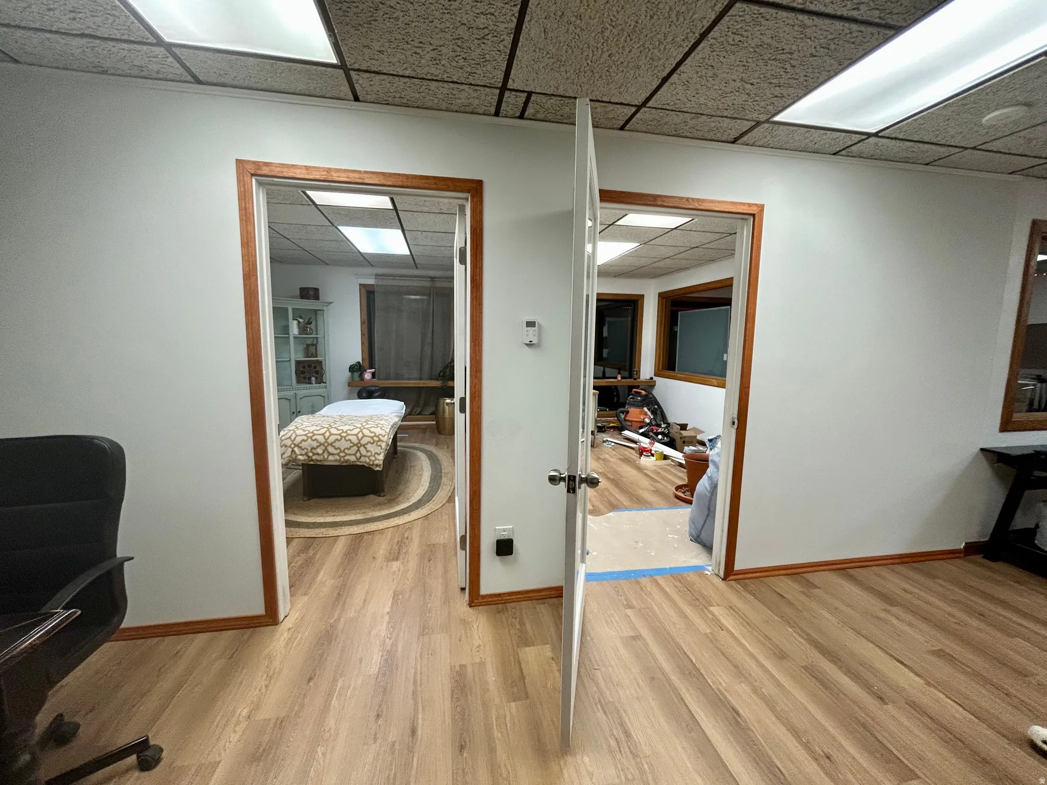 Interior space featuring wood-finish flooring throughout, two entryways with wood trim, white walls, and a suspended acoustic tile ceiling with integrated lighting