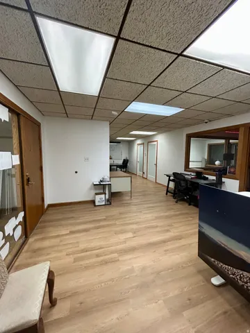 Spacious hallway featuring wood-finish flooring, white walls, and a suspended ceiling with integrated fluorescent lighting