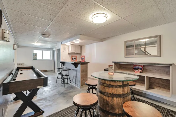 Finished basement featuring a built-in bar with light wood-finish cabinetry, a barrel-style high-top table with a glass surface, a shuffleboard table, and a built-in shelving unit