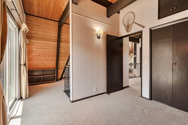 Carpeted interior space featuring a wood-paneled sloped ceiling, exposed beams, built-in shelving, a sliding glass door, and dark bi-fold closet doors