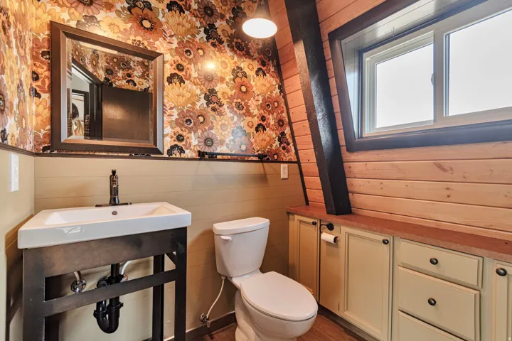 Bathroom with a modern vanity sink, dark framed mirror, floral wallpaper, wood-finish flooring, and a window with wood trim