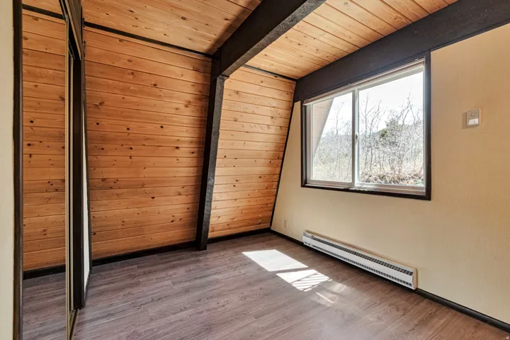 Room featuring wood-finish flooring, natural wood plank walls, exposed dark beams, and a large window overlooking trees