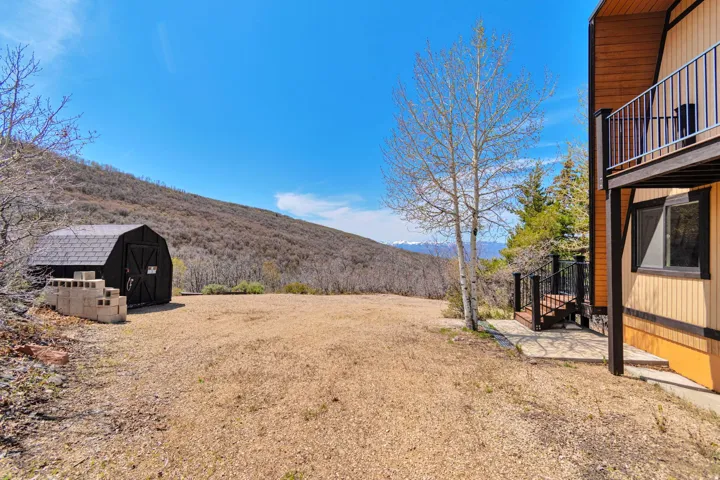 Detached storage shed with shingle roof, gravel yard, and wooded hillside backdrop