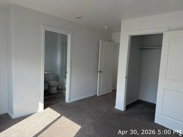 Carpeted room with white painted walls and trim, featuring a bifold closet with a metal hanging rod and a separate doorway leading to a bathroom with wood-finish flooring and a white toilet