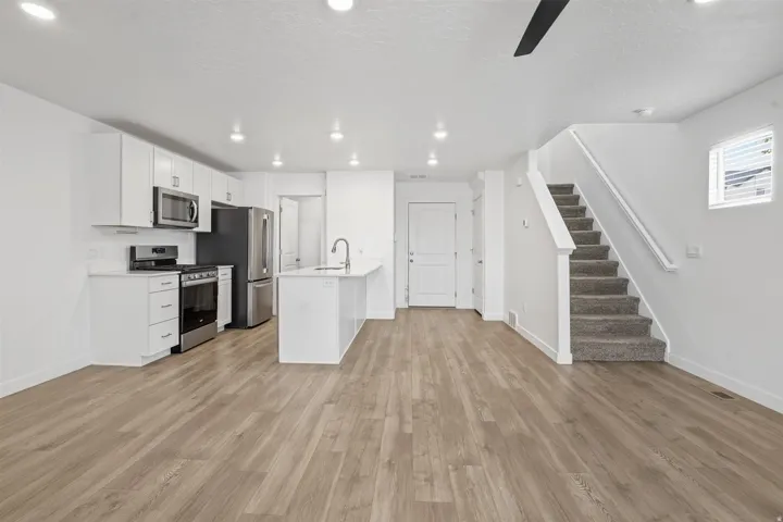 Open-concept living area featuring wood-finish flooring, a contemporary kitchen with white cabinetry, stainless steel appliances, a kitchen island with sink, recessed lighting, and a carpeted staircase with white handrails