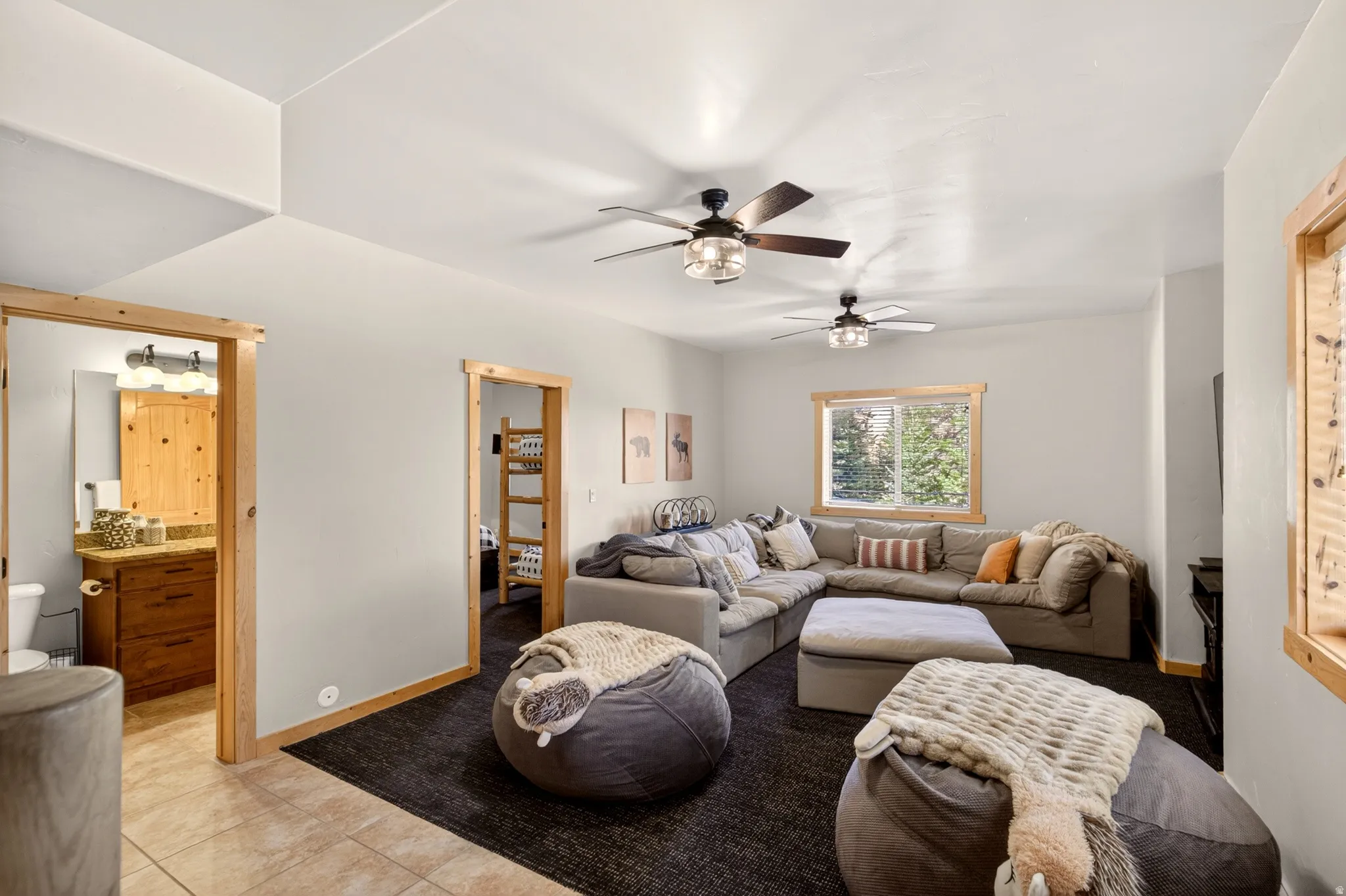 Spacious common area featuring light gray walls, two ceiling fans with integrated lighting, and a large window with natural wood trim