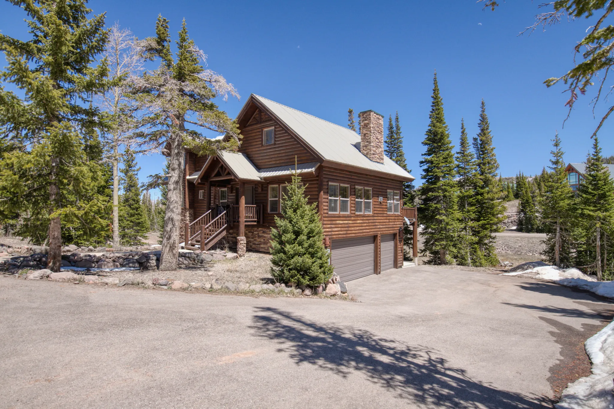 Log cabin exterior featuring a stone accent foundation, attached two-car garage, covered entry porch, and a prominent stone chimney