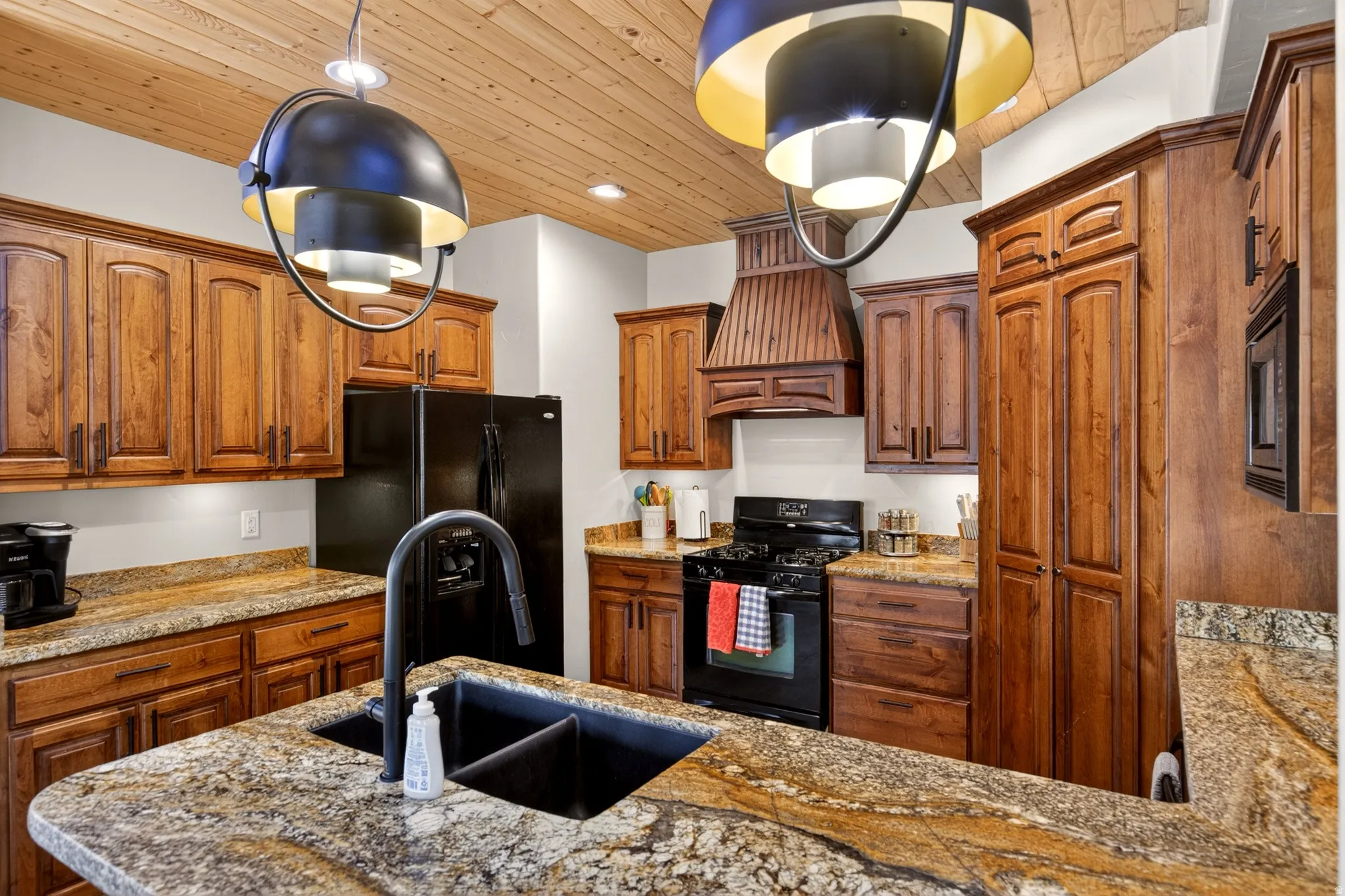 Kitchen featuring a wood-finish plank ceiling, granite countertops, wood cabinetry, a black appliance suite, and an island with an undermount sink