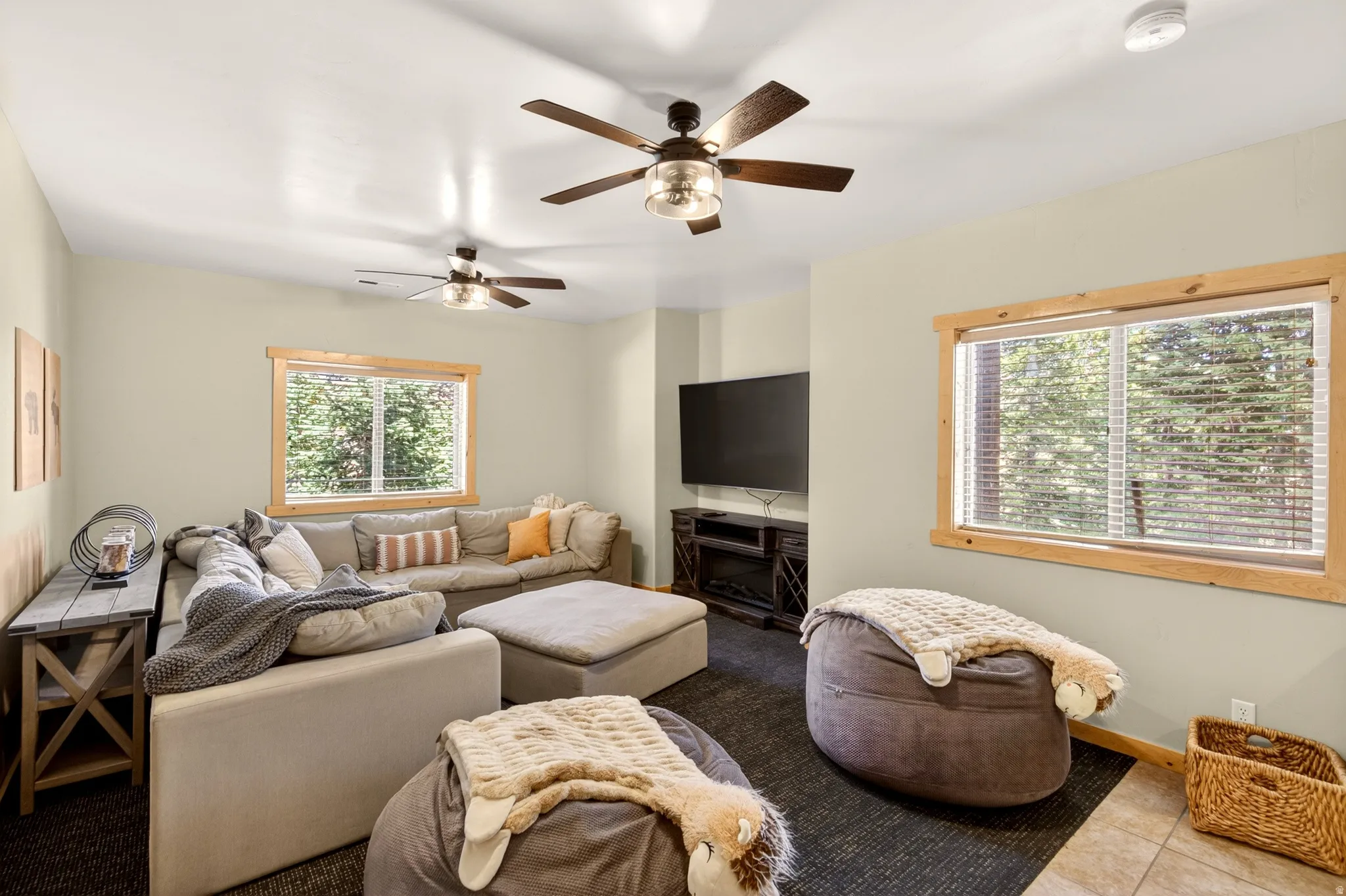 Spacious recreational room featuring two ceiling fans, wood-trimmed windows, dark carpeting, and a built-in electric fireplace media console