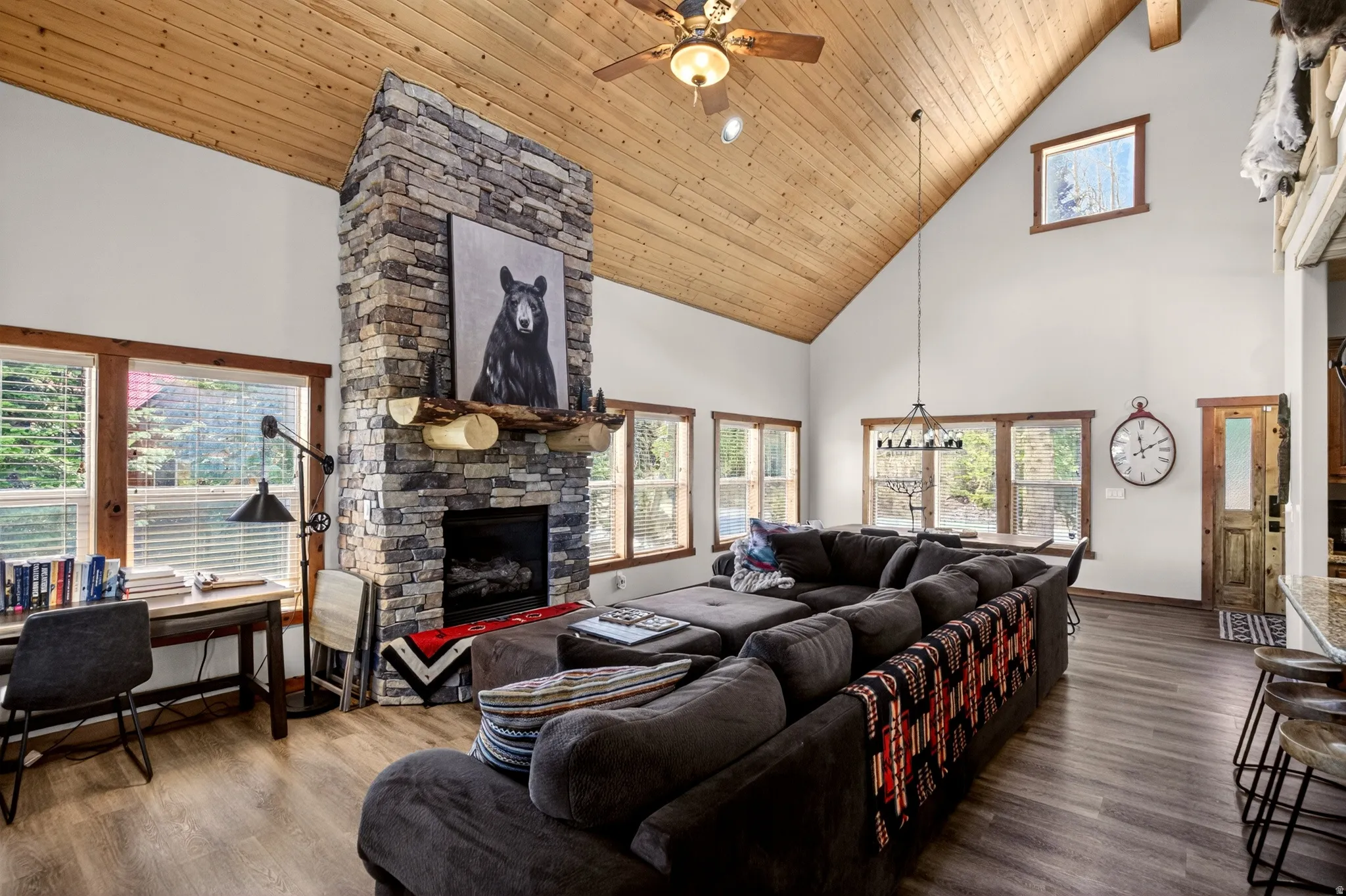 Spacious living area featuring a stone fireplace, vaulted wood-paneled ceilings, wood-finish flooring, and multiple windows with wood trim
