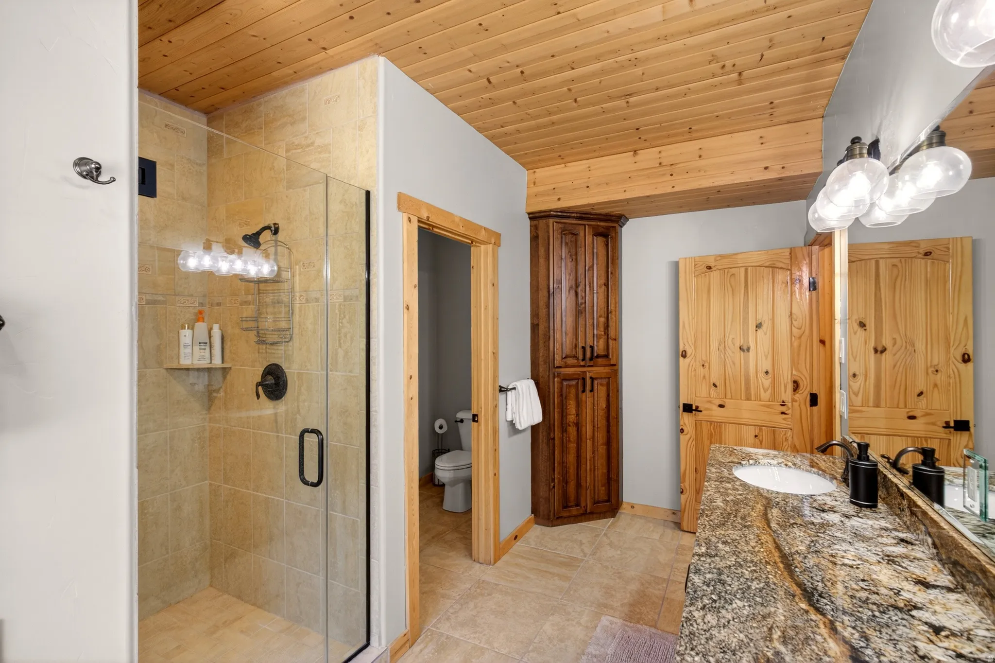 Bathroom featuring a wood-panel ceiling, glass-enclosed shower with tile surround, double vanity with a stone countertop, and custom wood cabinetry