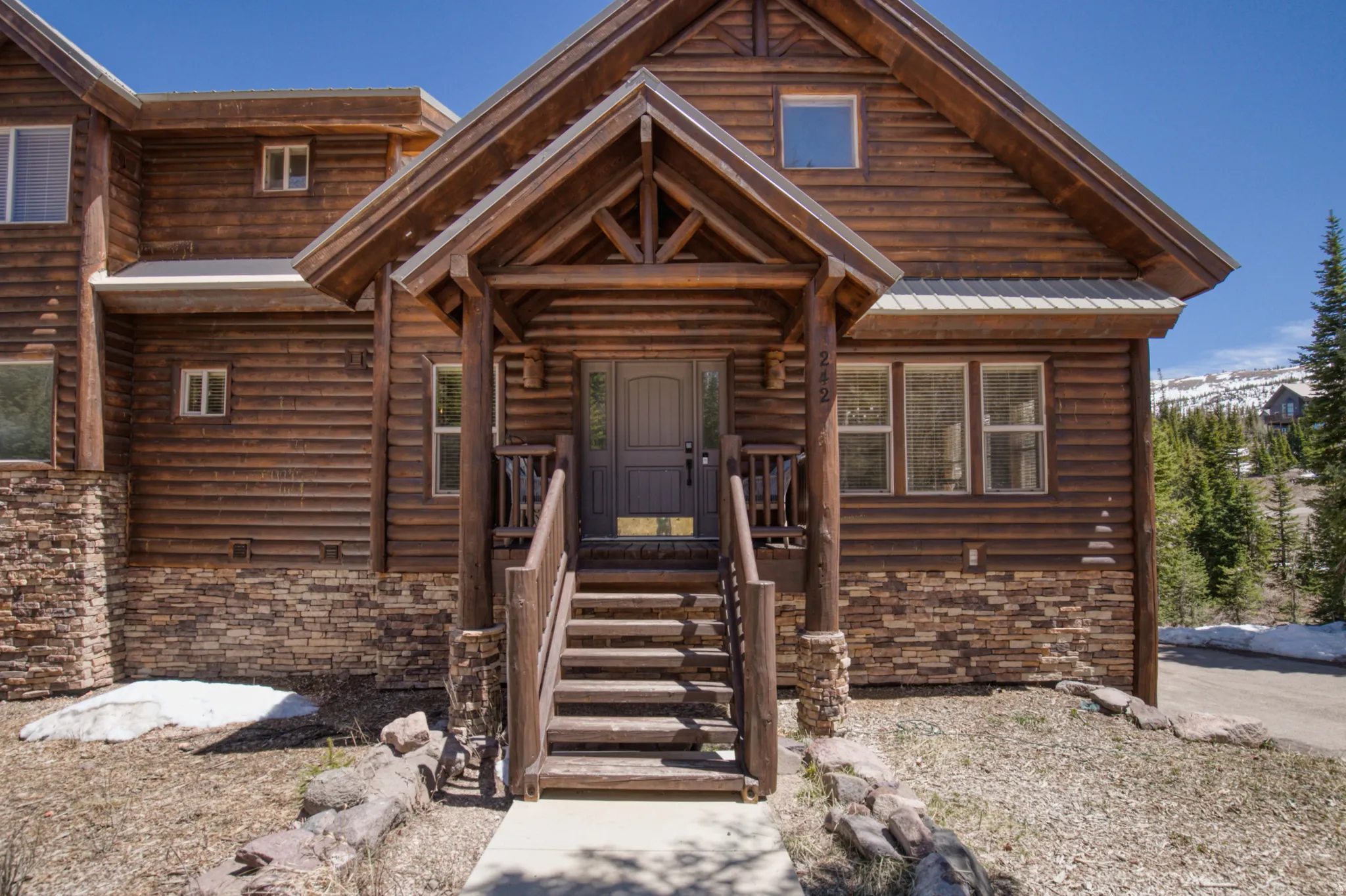 Log cabin exterior featuring a covered entry porch, visible natural wood construction, stone accent foundation, and multiple windows
