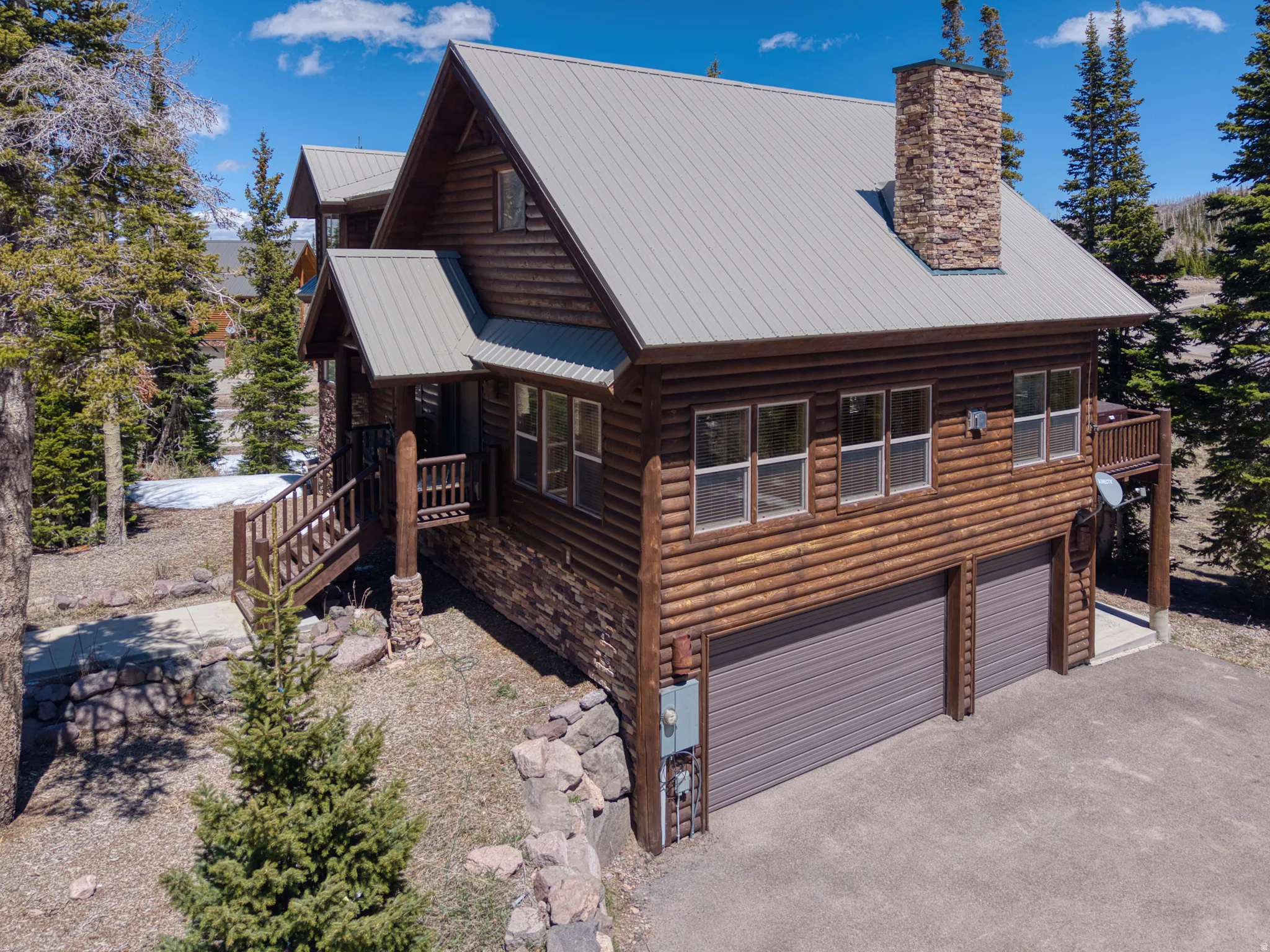 Log-sided home featuring a stone chimney, gray metal roof, and a two-car garage with brown doors