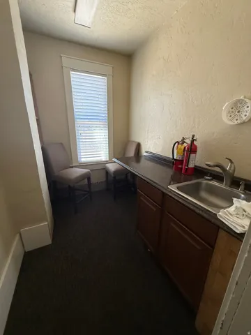 Compact utility area featuring a window with blinds, dark textured carpeting, baseboard trim, a countertop with an integrated sink, and wood-finish cabinetry