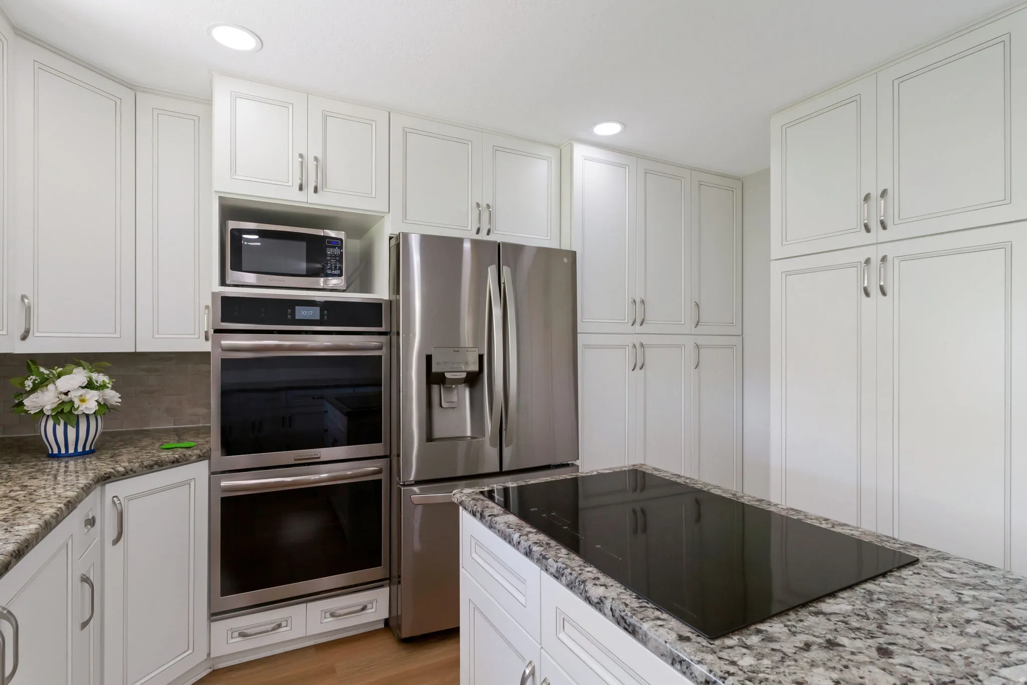 Kitchen featuring extensive white cabinetry, granite countertops, built-in double ovens, a stainless steel refrigerator, and an island cooktop