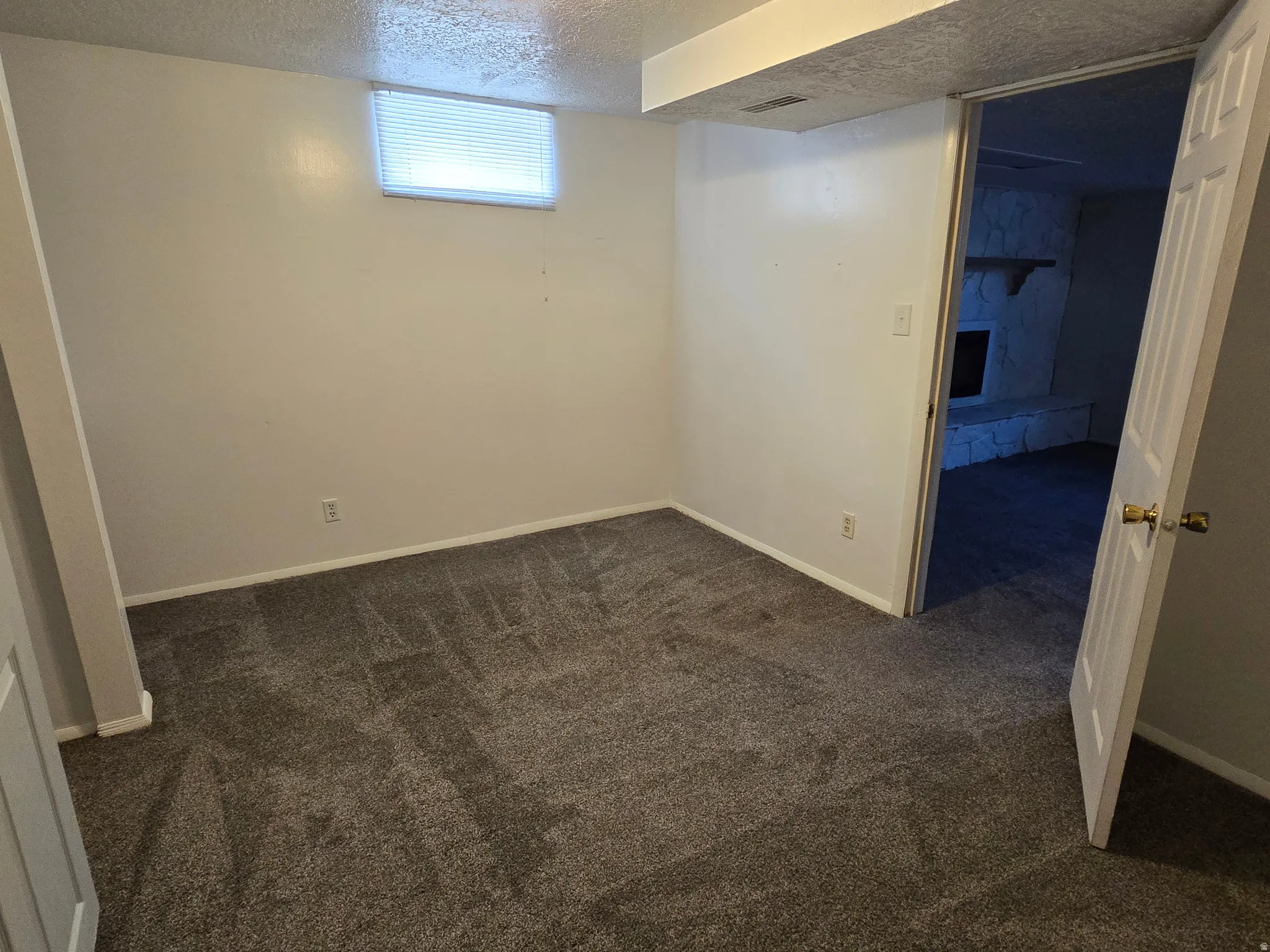 Carpeted bedroom with a single window, white walls, and a visible doorway leading to an additional space featuring a stone fireplace