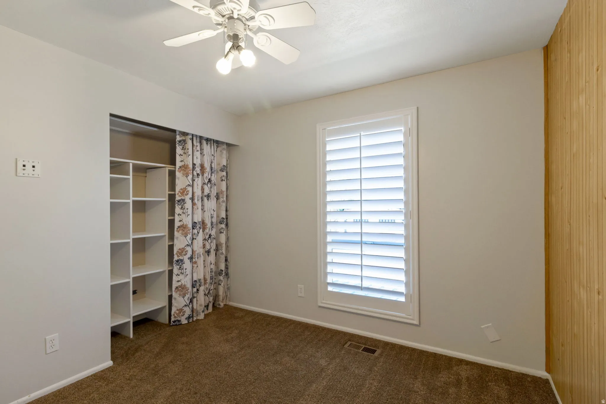 Carpeted room featuring a ceiling fan with integrated lighting, a window with white plantation shutters, and built-in shelving in closet