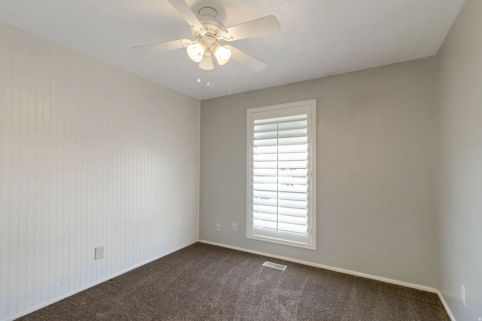 Neutral-toned bedroom featuring a white ceiling fan with integrated lighting, a window with plantation shutters, vertical plank wall paneling, and carpet flooring