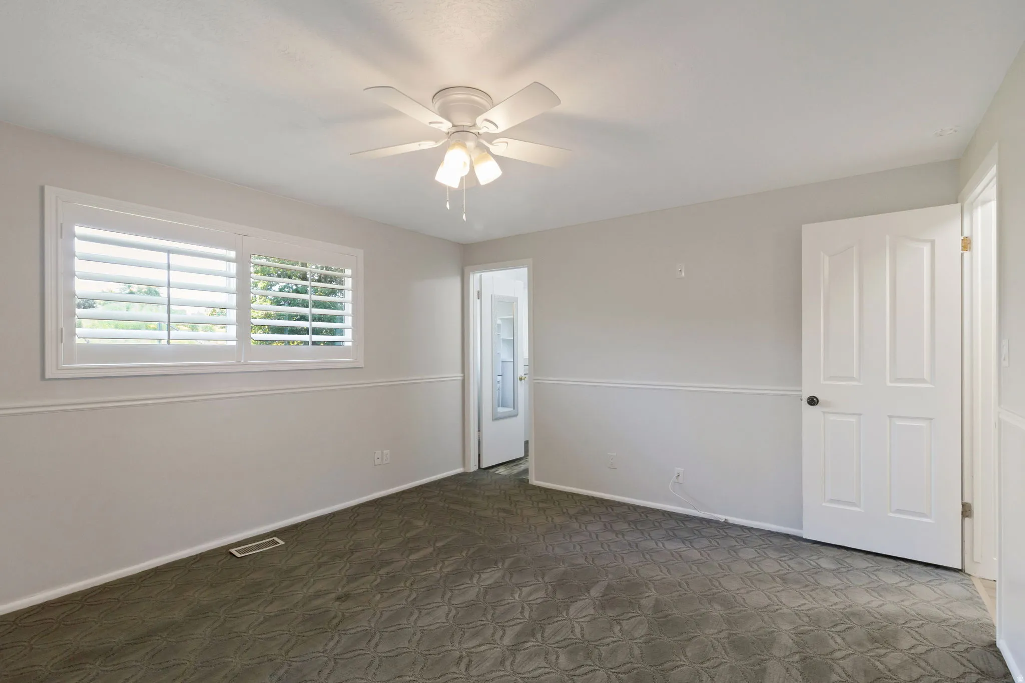 Interior bedroom featuring a white ceiling fan with integrated lighting, window with white plantation shutters, gray patterned carpeting, and white baseboards with chair rail molding
