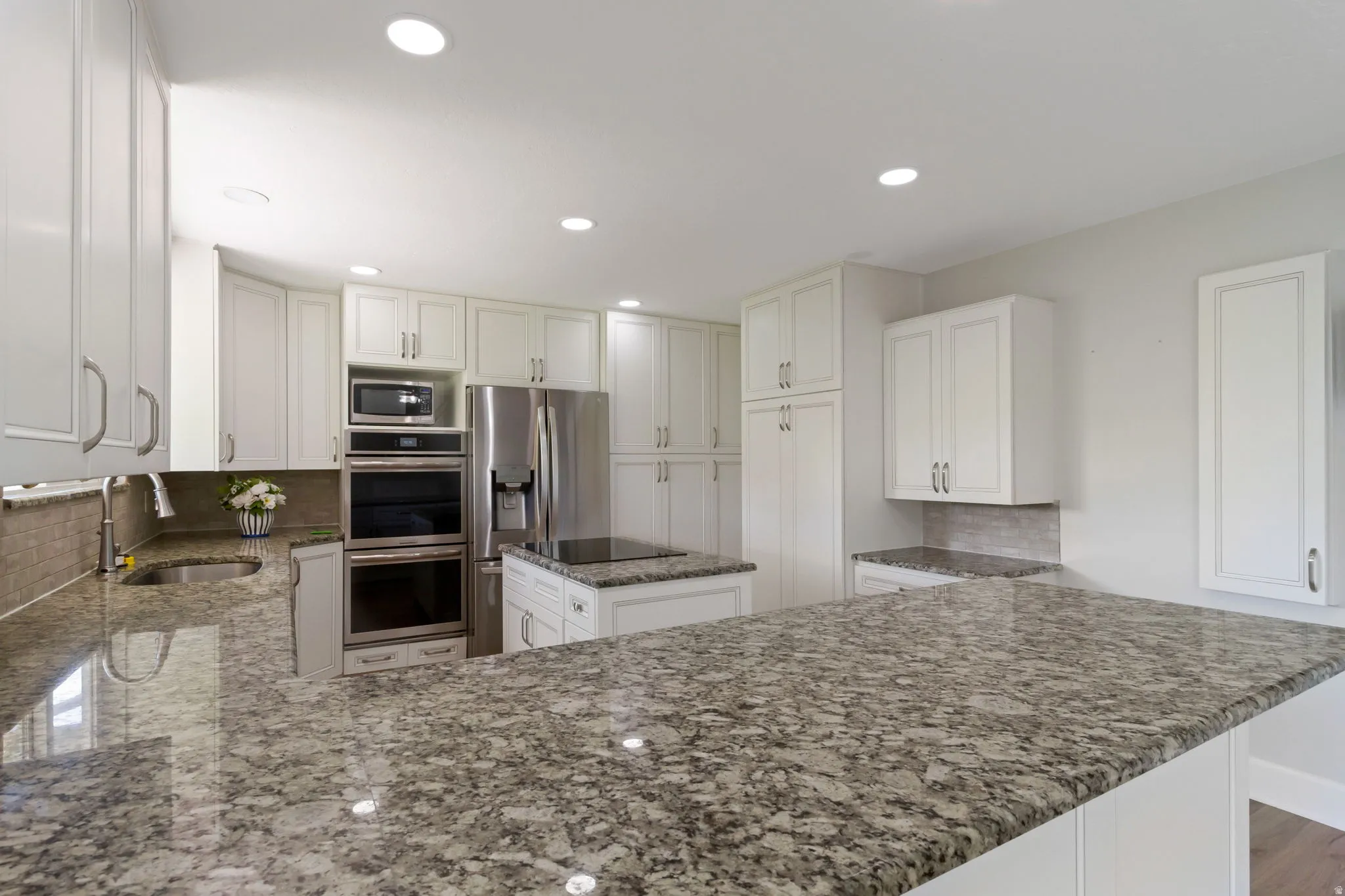 Spacious kitchen featuring extensive granite countertops, white cabinetry with brushed nickel hardware, stainless steel appliances including a built-in microwave and double ovens, and a tile backsplash