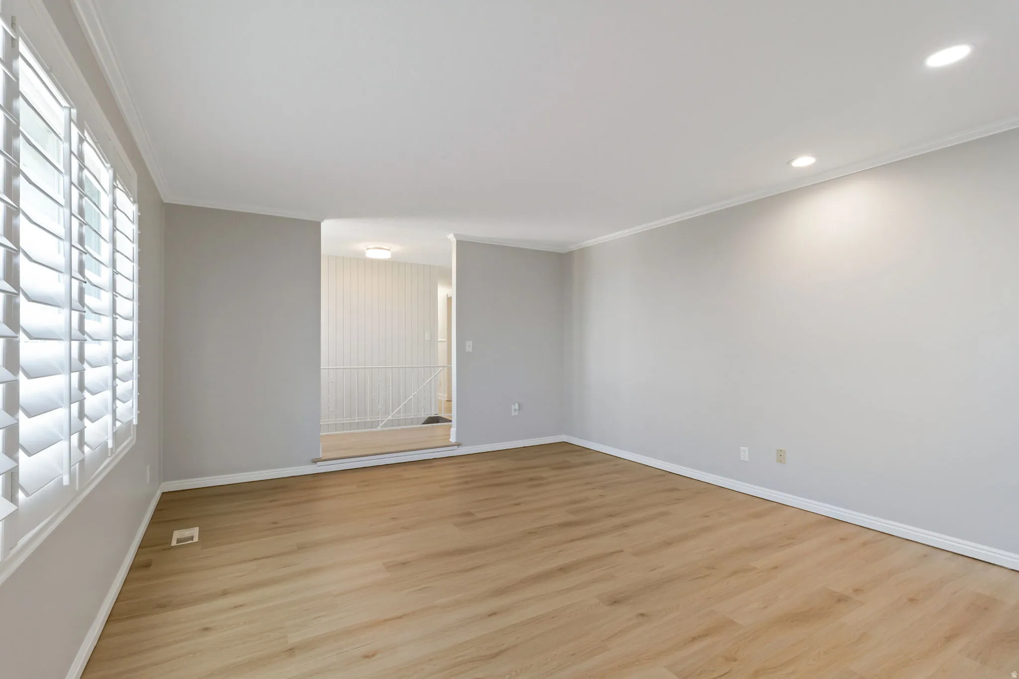 Spacious room featuring light wood-look flooring, plantation shutters, recessed lighting, crown molding, and neutral wall tones