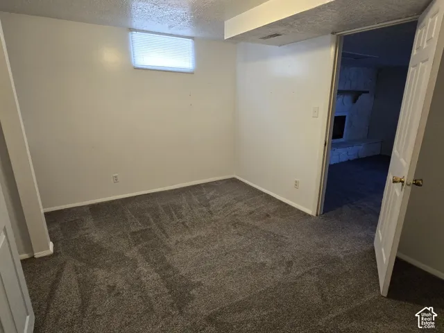 Carpeted bedroom with a single window, white walls, and a visible doorway leading to an additional space featuring a stone fireplace