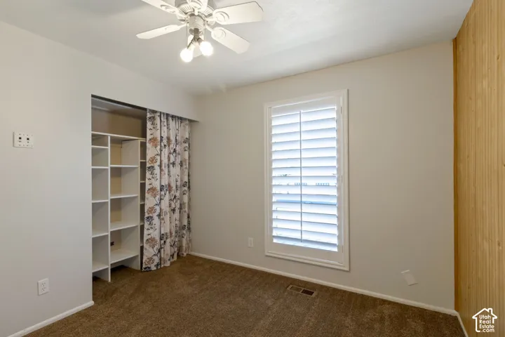 Carpeted room featuring a ceiling fan with integrated lighting, a window with white plantation shutters, and built-in shelving in closet