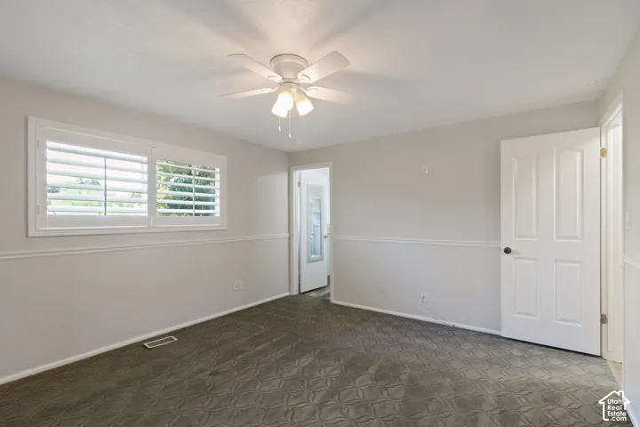 Interior bedroom featuring a white ceiling fan with integrated lighting, window with white plantation shutters, gray patterned carpeting, and white baseboards with chair rail molding
