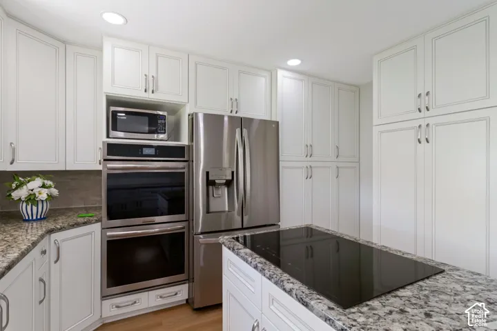 Kitchen featuring extensive white cabinetry, granite countertops, built-in double ovens, a stainless steel refrigerator, and an island cooktop