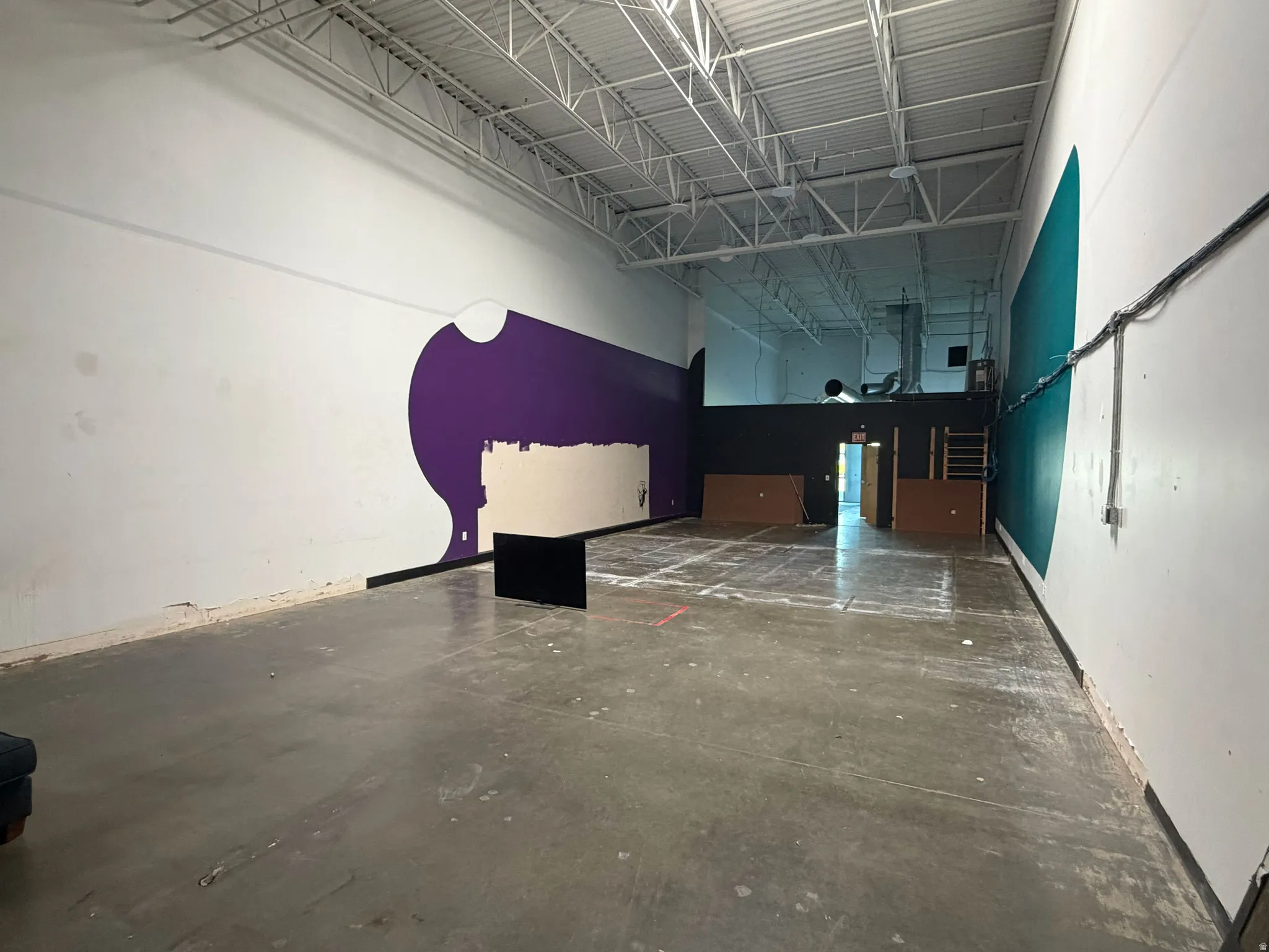 Expansive commercial space featuring exposed ceiling joists, industrial-style concrete flooring, a partial mezzanine level, and a combination of white, purple, and teal wall finishes