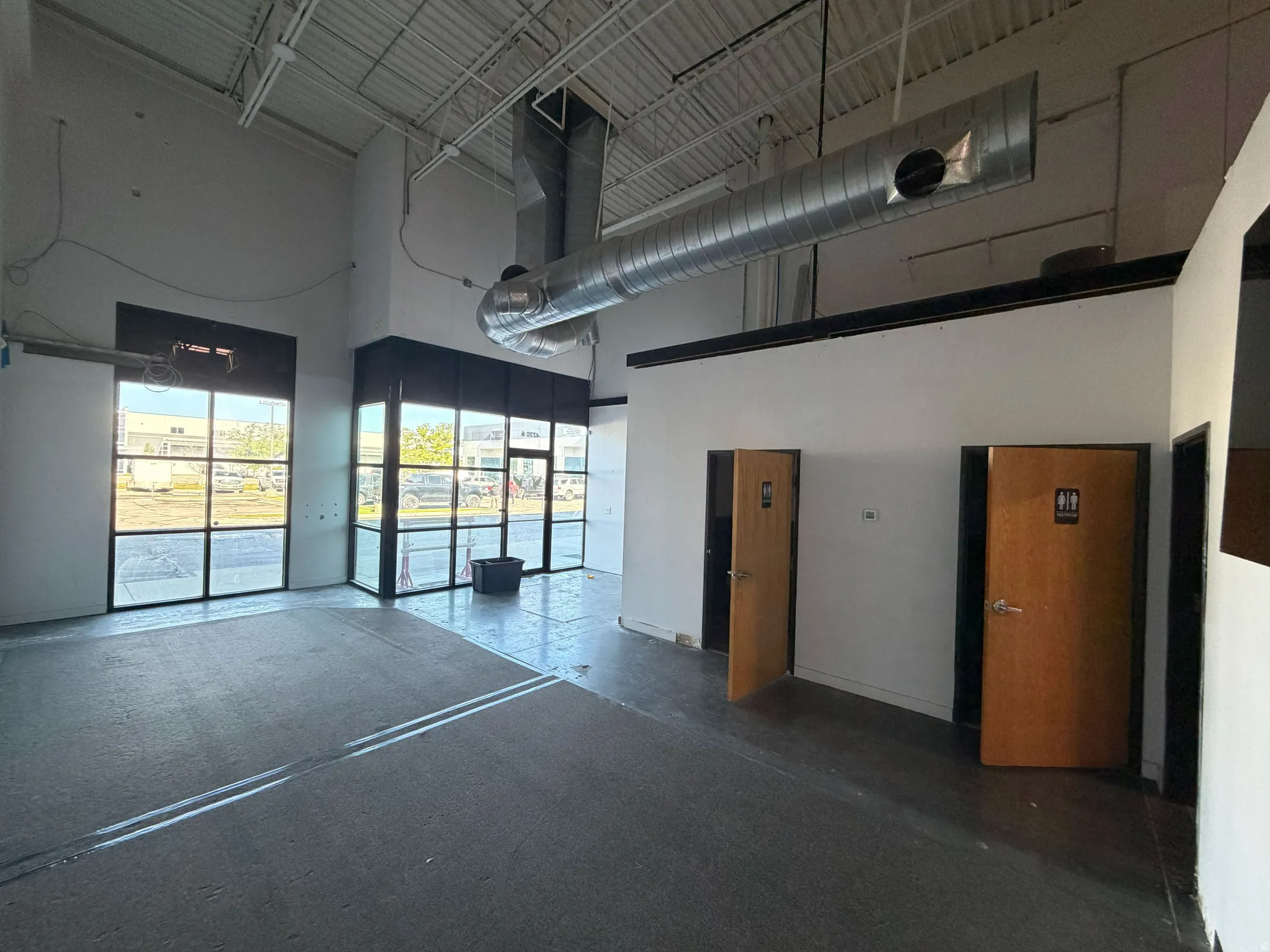 Spacious commercial interior featuring concrete flooring, high ceilings with exposed ductwork, and large storefront windows with black framing