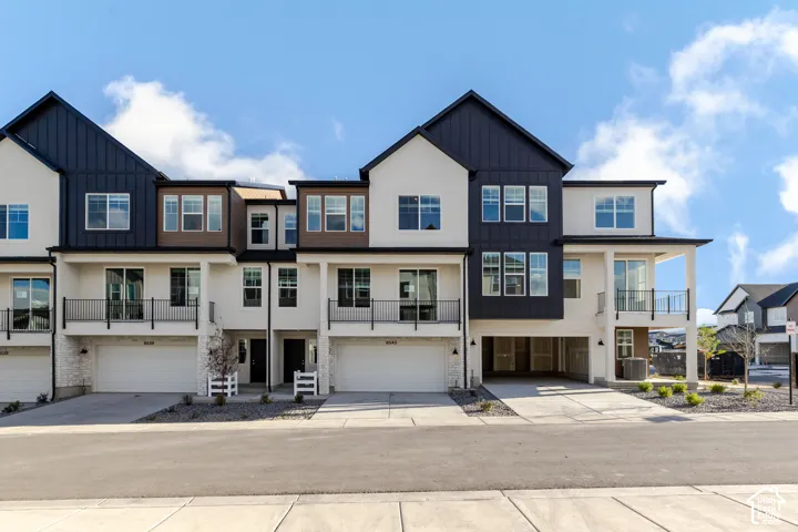 Multi-story townhome exterior featuring stucco and metal panel siding, attached garages, and second-story metal-railed balconies