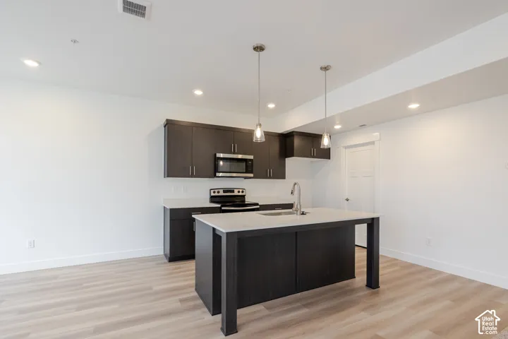 Kitchen featuring dark-toned cabinetry, light-toned countertops, wood-finish flooring, a central island with an integrated sink, and dual glass pendant lighting