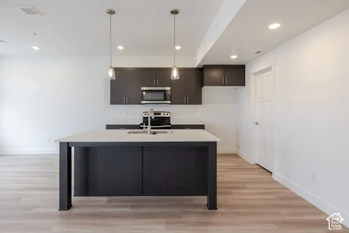 Kitchen featuring dark-toned cabinetry, light-colored countertops, center island with integrated stainless steel sink, wood-finish flooring, and dual glass pendant lighting