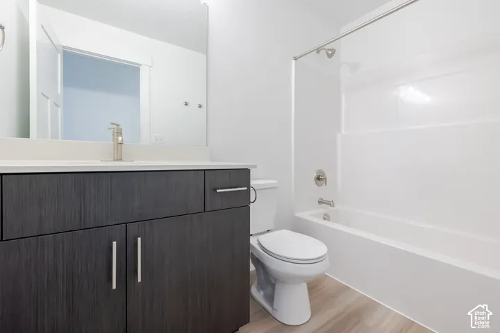 Dark-toned cabinetry with modern hardware, white vanity countertop, wall-mounted mirror, wood-finish flooring, and a combined tub and shower enclosure with a stainless steel faucet