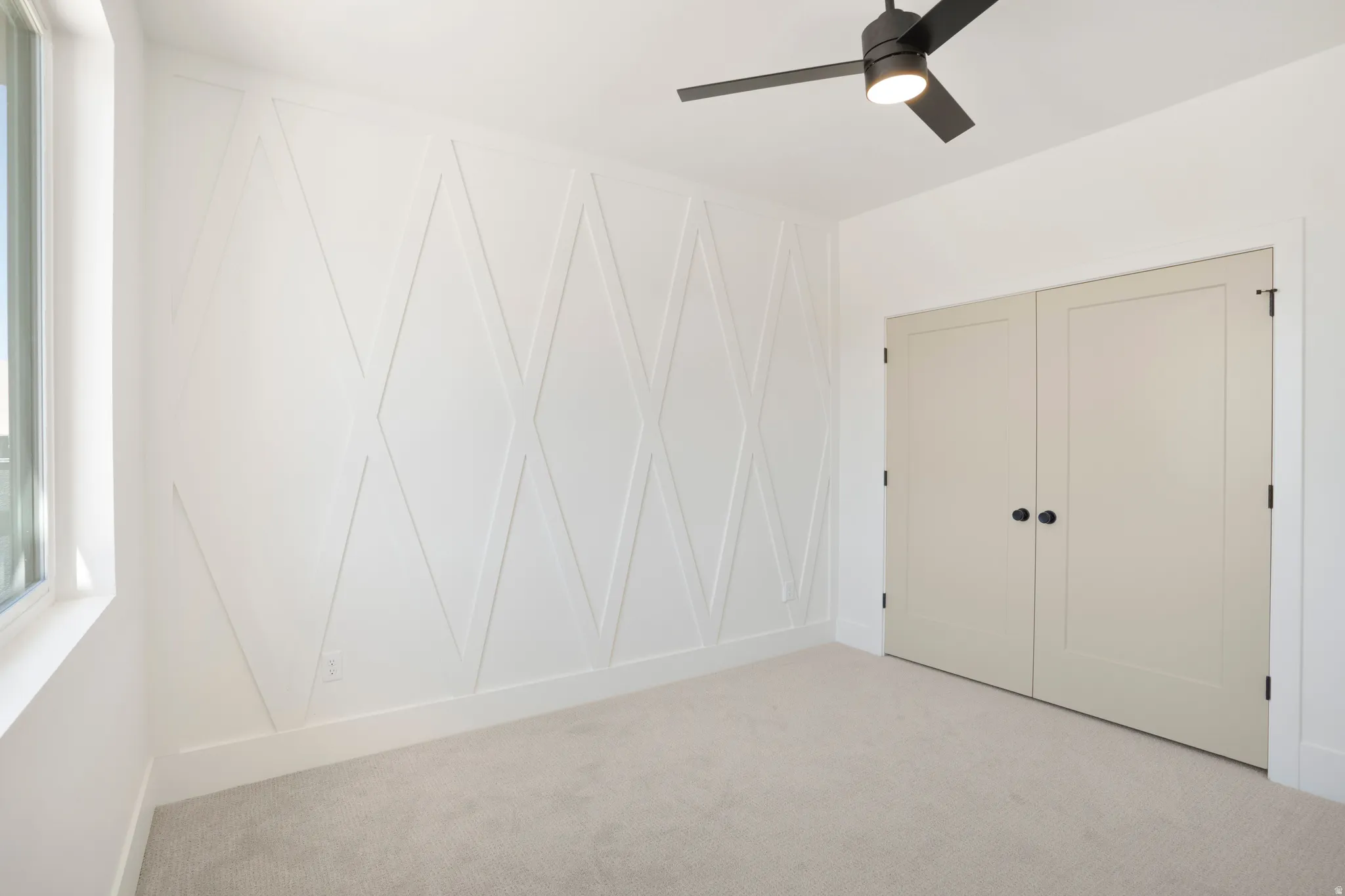 Room featuring a decorative accent wall with geometric paneling, light-colored carpeting, and a black ceiling fan with integrated lighting