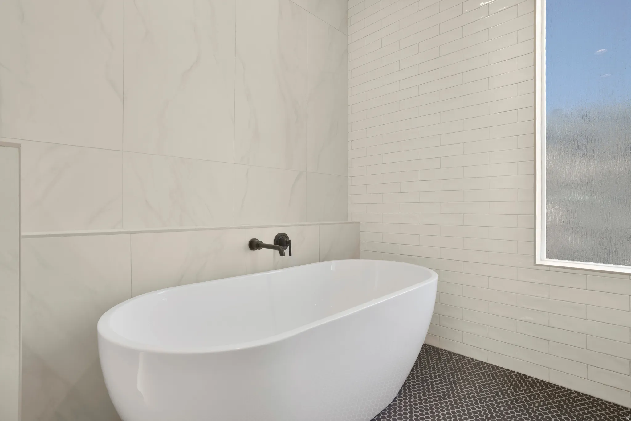Freestanding soaking tub with a wall-mounted matte black faucet, set against large format wall tiles and subway tile accents