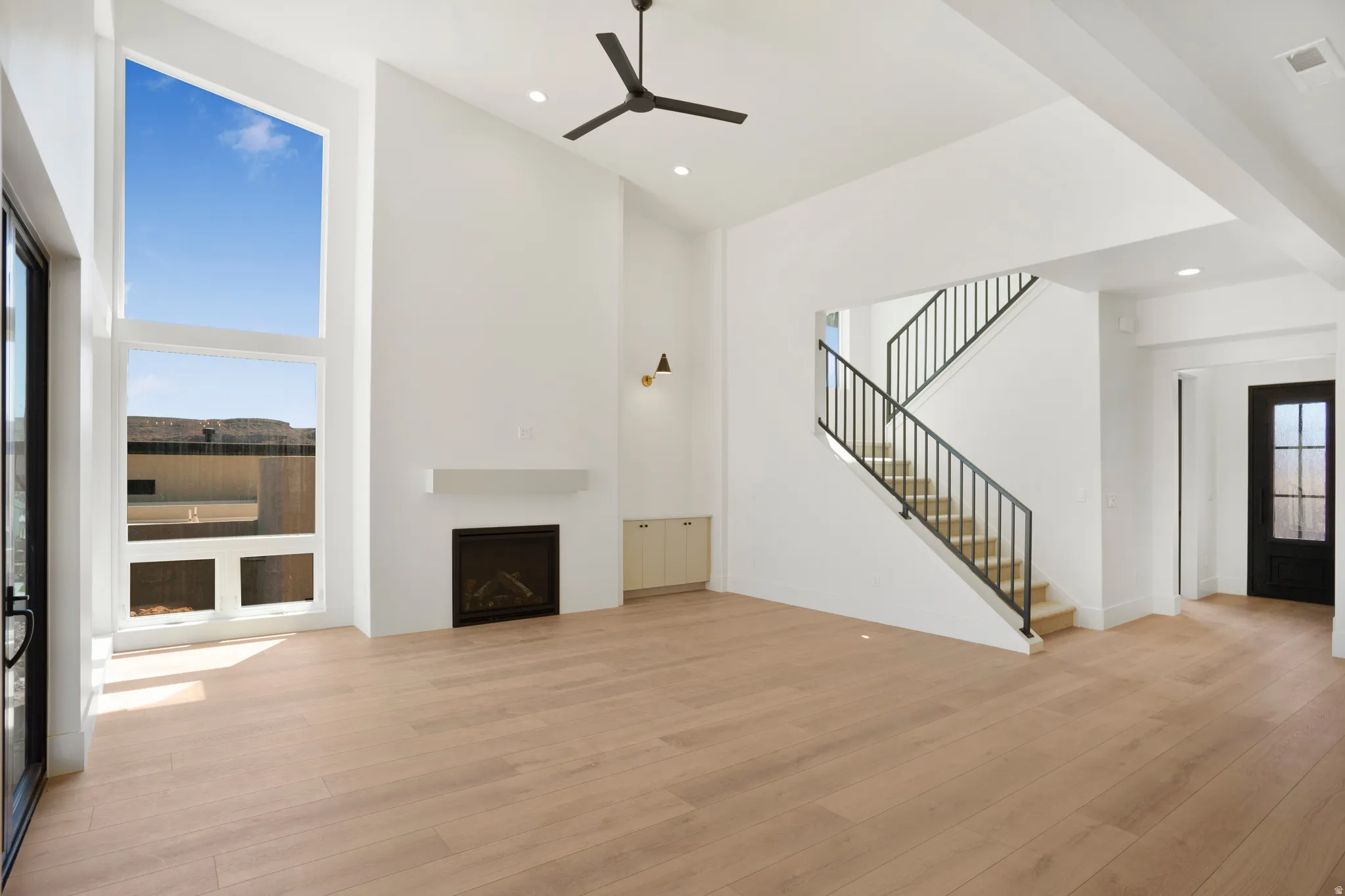 Vaulted ceiling with a contemporary ceiling fan, prominent fireplace with a clean-line mantel, large windows, light wood-finish flooring, and an open staircase with a metal railing