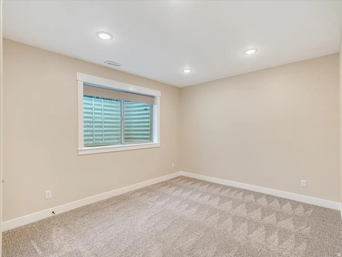 Spacious room featuring neutral carpeting with a subtle geometric pattern, recessed lighting, and a window with a roller shade
