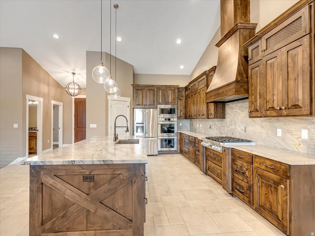 Spacious kitchen featuring an extended center island with integrated sink, wood-finish cabinetry, stainless steel appliances, a large range hood, and tile flooring