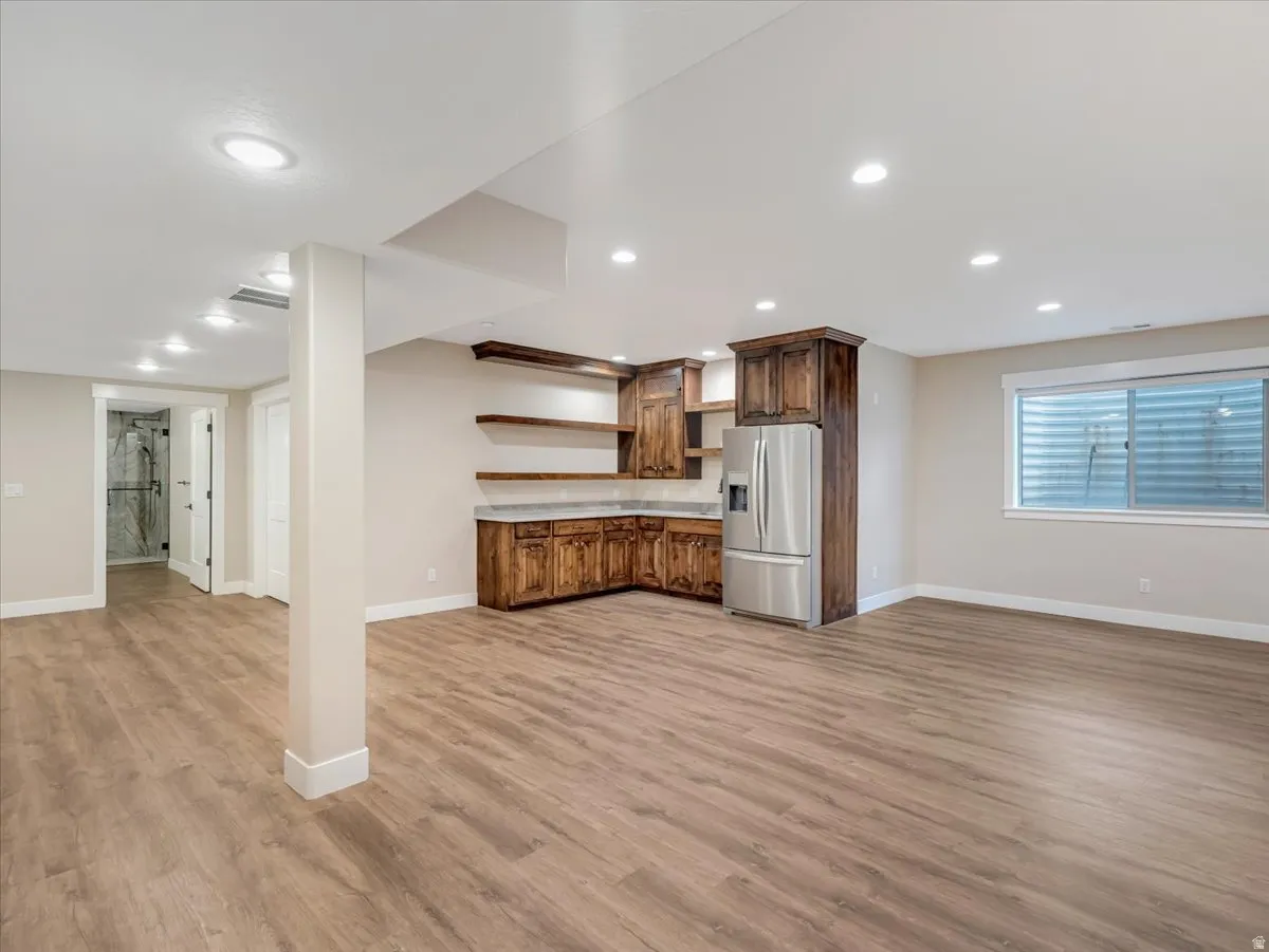 Spacious room featuring wood-finish flooring, recessed lighting, a built-in wet bar with wood cabinetry and open shelving, a stainless steel refrigerator, and a basement window