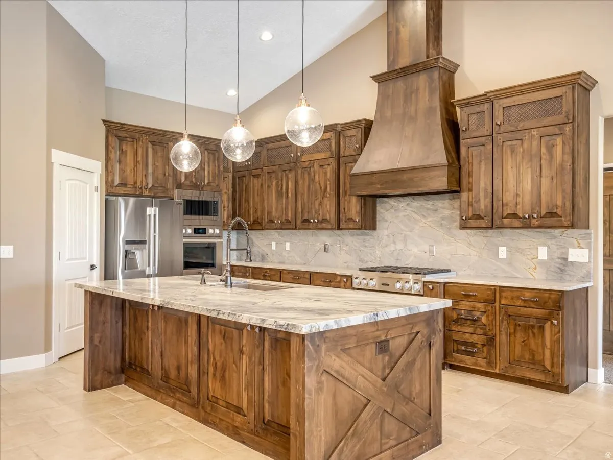 Spacious kitchen featuring an expansive central island with a farmhouse-style accent, abundant wood-finish cabinetry, stainless steel appliances, a professional-grade range, and elegant stone countertops with a coordinating full-height backsplash